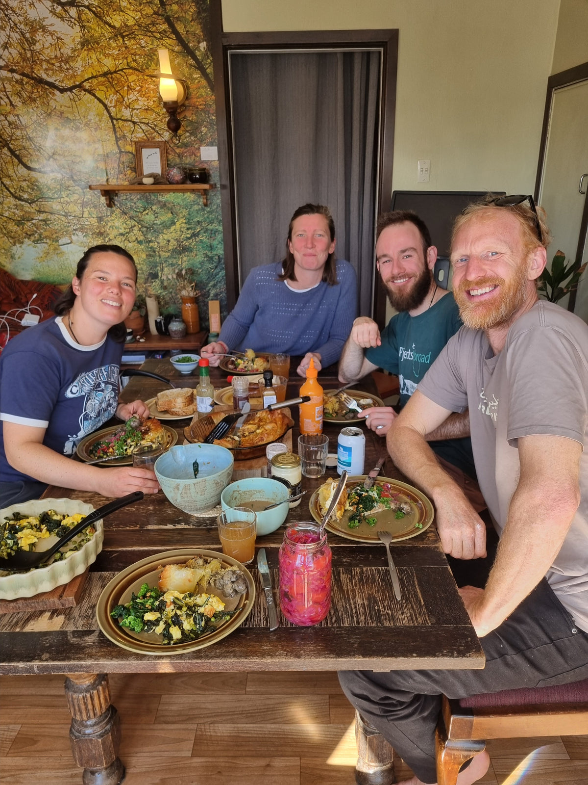 Four people sitting together around a rustic wooden dining table laden with food, bowls, and drinks in a home interior with a large tree mural on the wall.