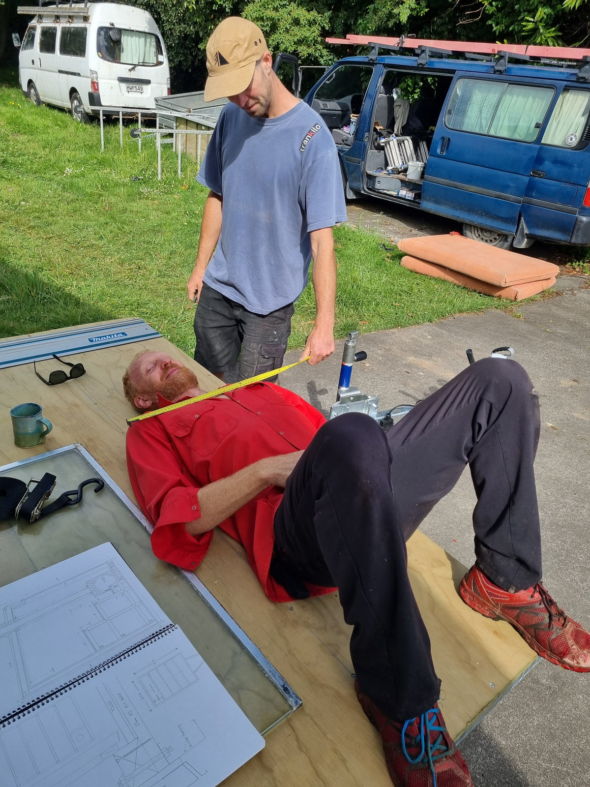 A man in a blue shirt and tan cap measures the width of another man lying on a wooden board with a tape measure, outdoors near vans and a grassy area.
