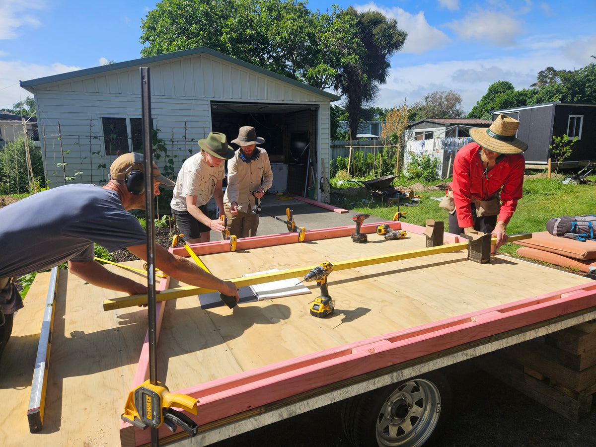 Several people working outdoors on a wooden trailer frame, with tools and lumber visible in a backyard setting with a shed and trees in the background.