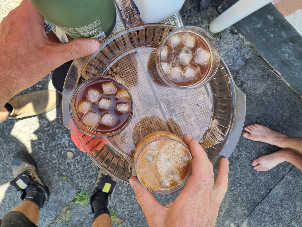 Overhead view of a round wooden tray holding three glasses of iced coffee or latte with ice, held by hands outdoors on a paved surface. A green bottle is partially visible at the top left.