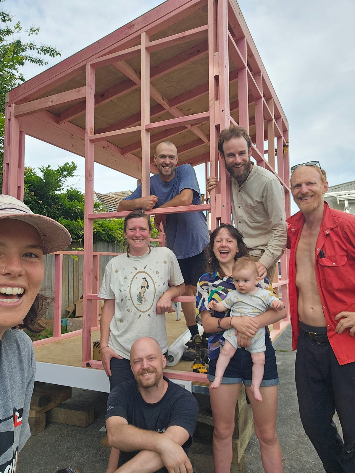 A group of seven people posing together in front of a large pink wooden house frame structure outdoors on a cloudy day.