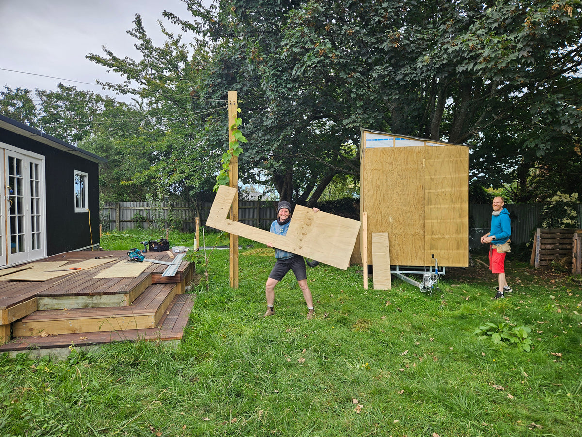 Two people outdoors in a backyard setting, working on a wooden structure. One person is carrying a large plywood panel, while another stands nearby next to a partially assembled wooden wall panel on a wheeled dolly.