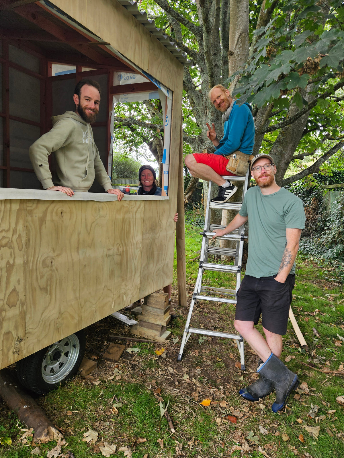 Four people are gathered outdoors next to a mobile wooden structure on wheels, likely a tiny house or workshop, in a grassy wooded area. One person stands on a metal ladder near the front of the structure, while others look on from windows and the side.
