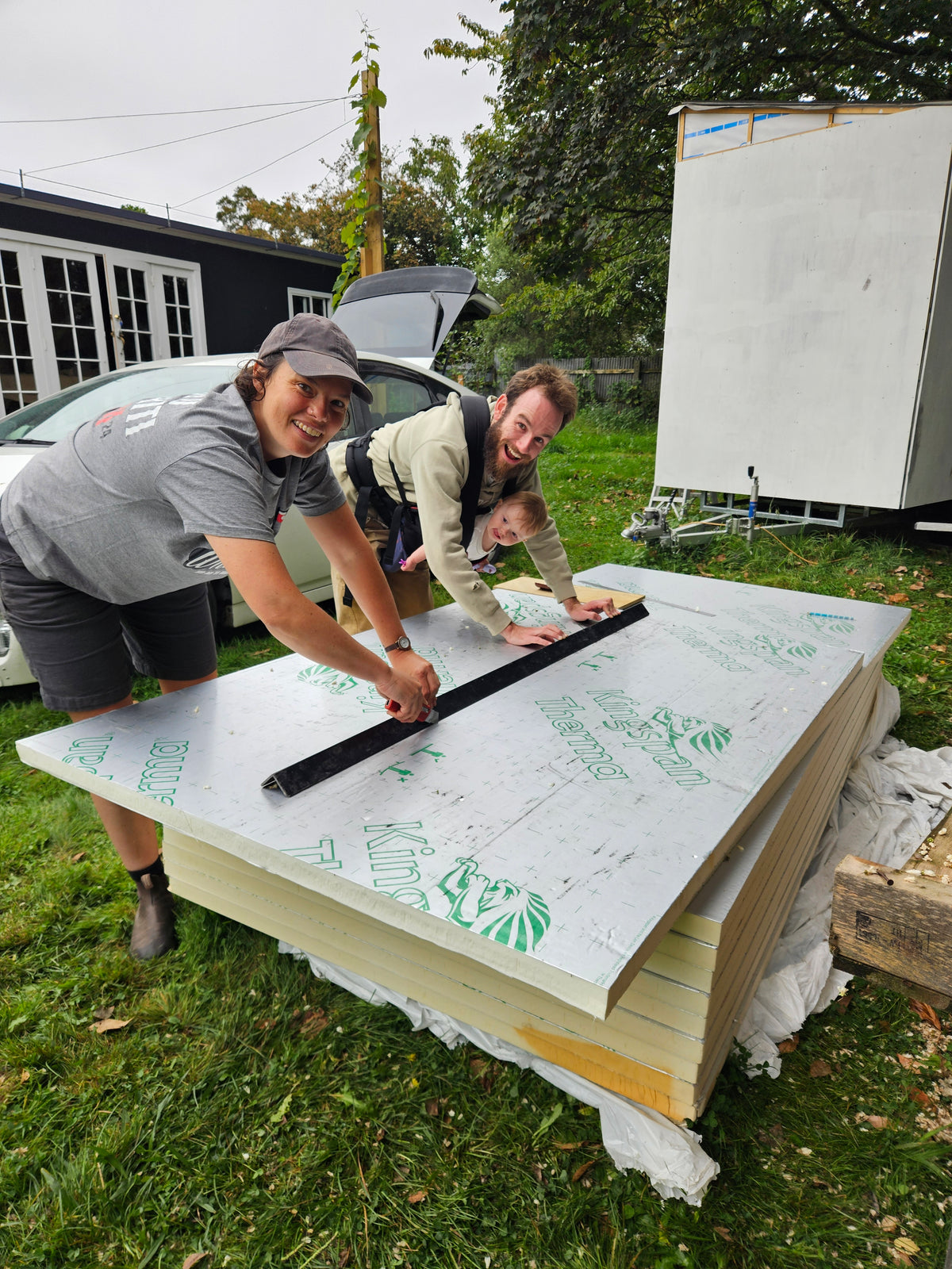 Two people outdoors on grass measuring a large stack of white and yellow insulation boards with a long black ruler. A house, car, and white trailer are visible in the background.