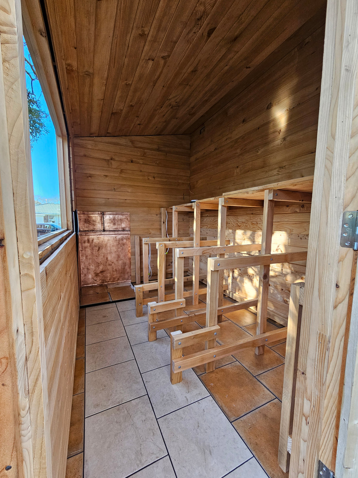 A rustic wooden cabin interior with unfinished bunk beds made from light wood frames, tiled floor, and a window showing blue sky.