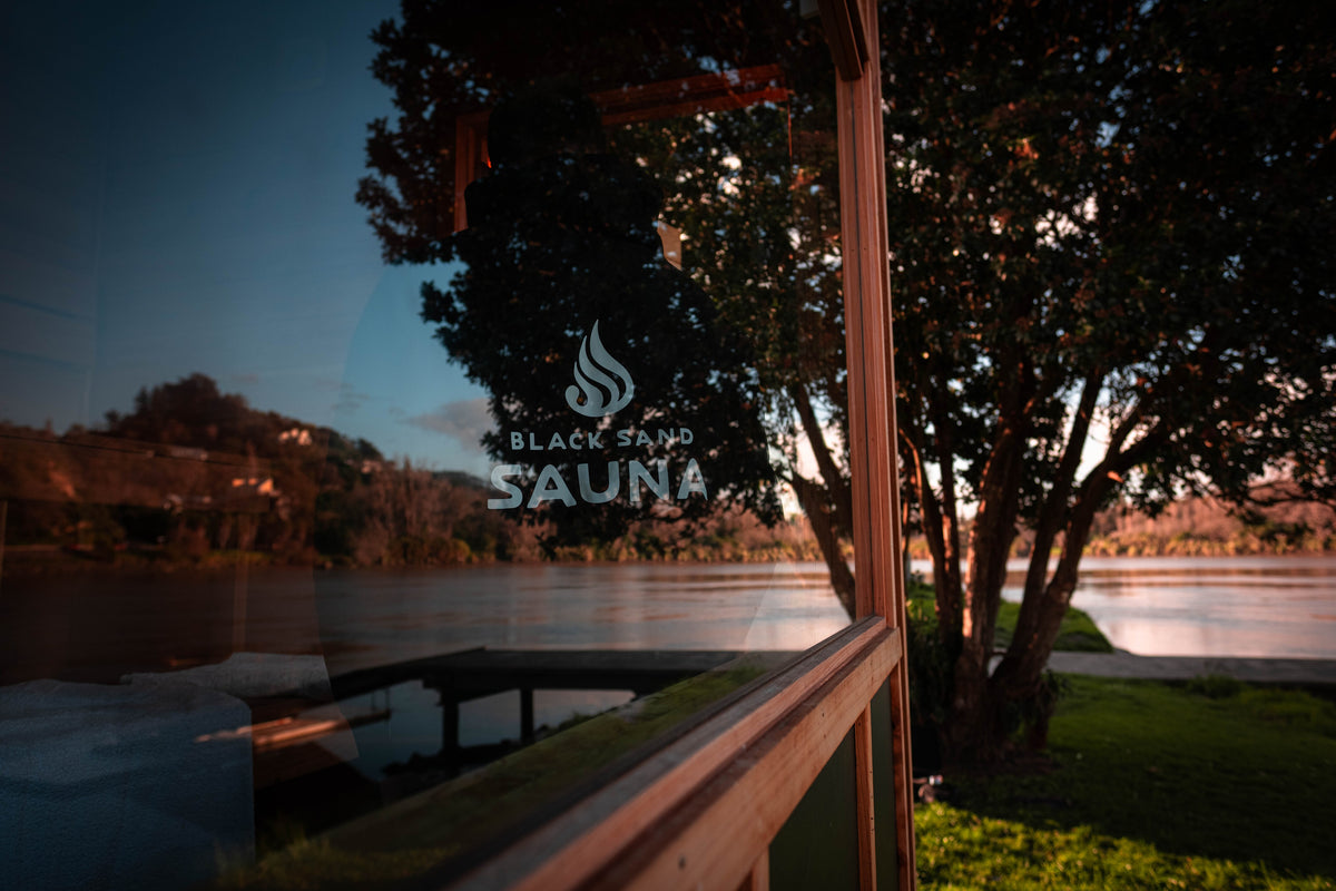 A scenic outdoor view of a river and trees reflected in a glass window, with the words 'BLACK SAND SAUNA' printed on the glass.