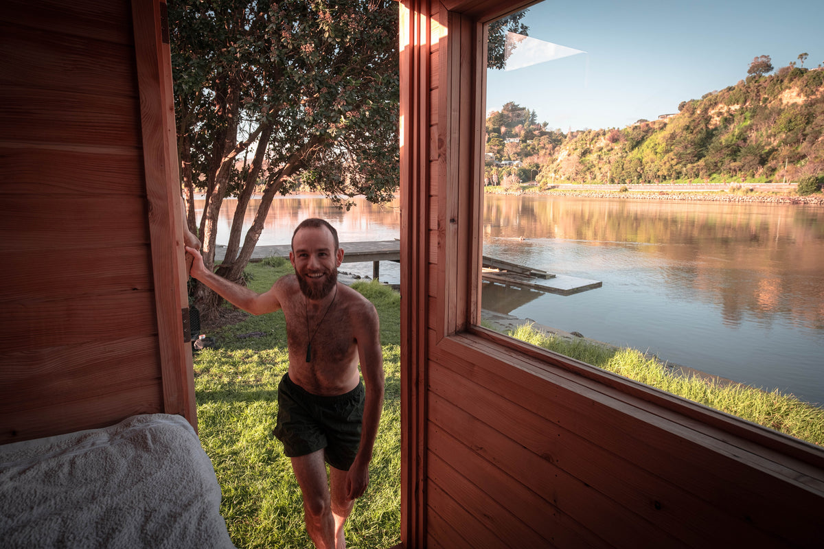 A shirtless man with a beard wearing entering a sauna, a window looking out toward a scenic river and forested hillside during golden hour.