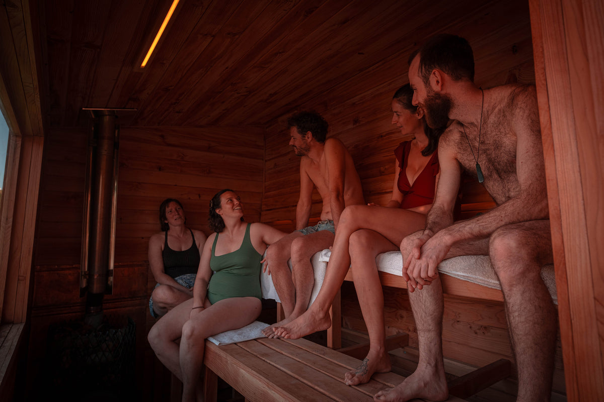 Several people sitting on wooden benches inside a wood-paneled sauna room with a visible stove and warm ambient lighting.