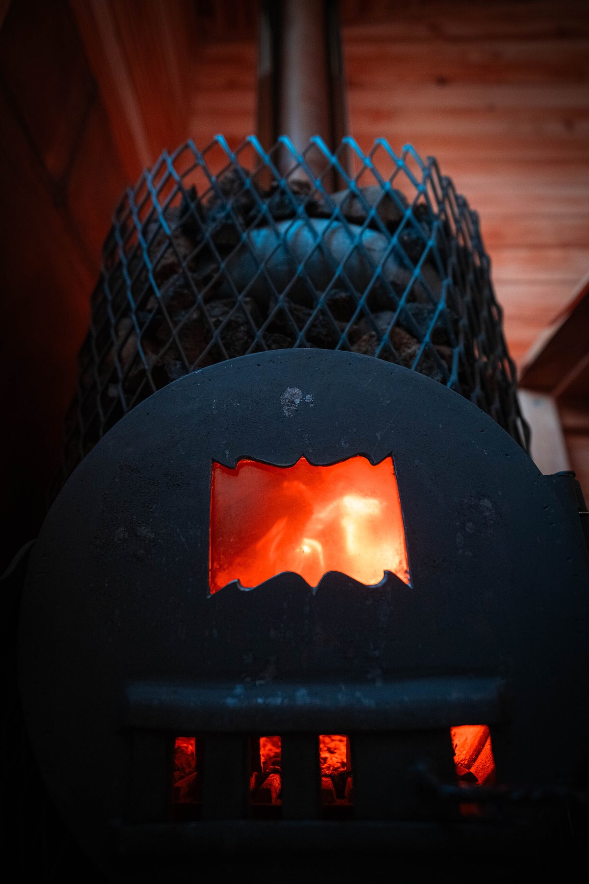 A close-up shot of a black metal wood-burning stove with glowing orange and red flames visible through the front window and bottom vents, set against a dark background with a blue wire mesh guard.
