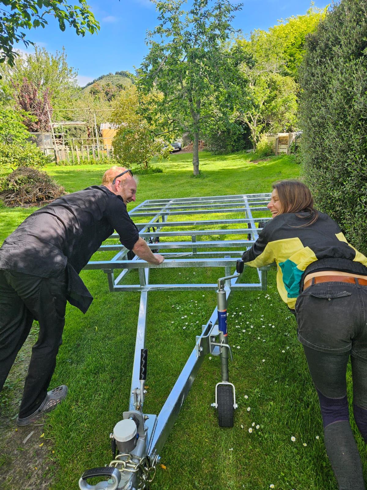 A man and a woman outdoors on a grassy lawn assembling a silver metal trailer frame with a single wheel and hitch mechanism.
