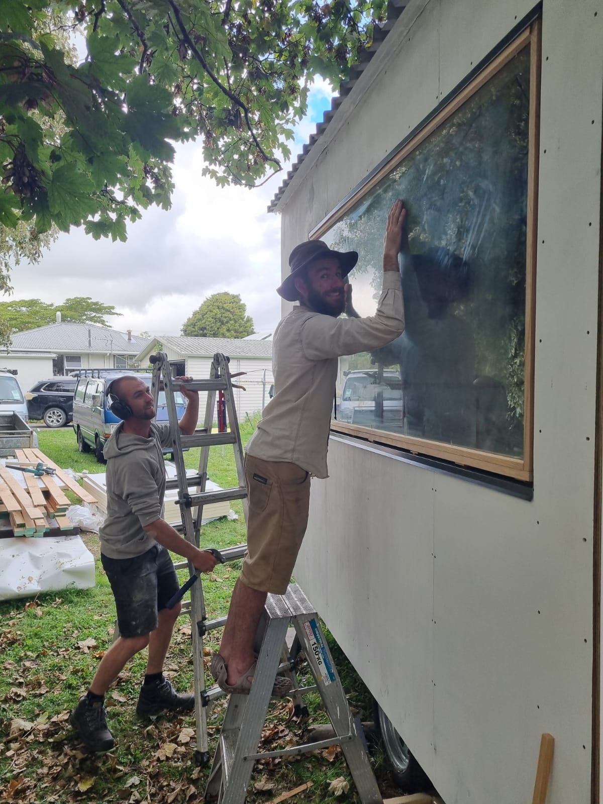 Two men installing a large rectangular window on the side of a white modular home or trailer outdoors, with ladders, trees, and construction materials visible in the scene.