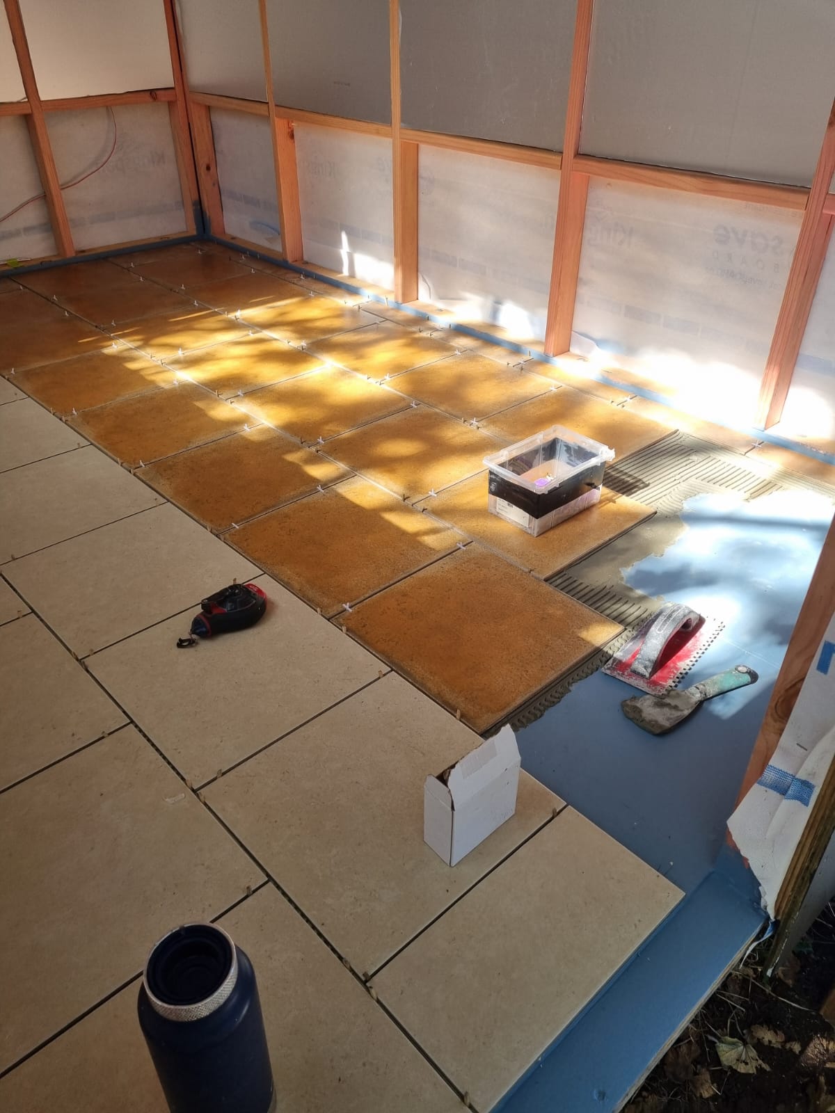 A room under construction or renovation with partially installed square floor tiles in beige and brown tones, exposed wooden framing, a blue base layer, and construction tools on the floor.