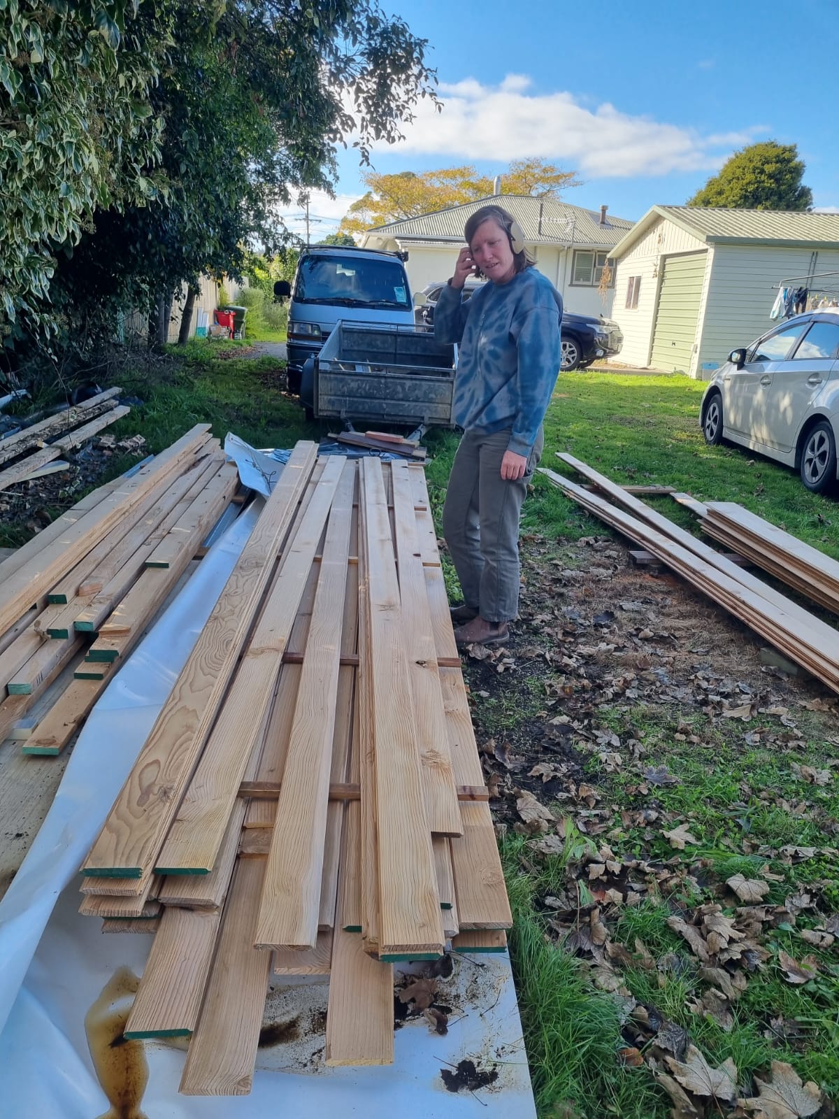 A person standing outdoors in a backyard or workshop area next to a large stack of raw lumber planks on a white sheet, with a truck, house, and car in the background.