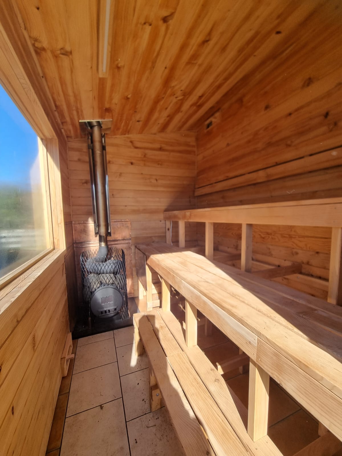A wooden sauna interior with tiered wooden benches, a wood-paneled ceiling and walls, a metal chimney pipe, and a window on the left side.