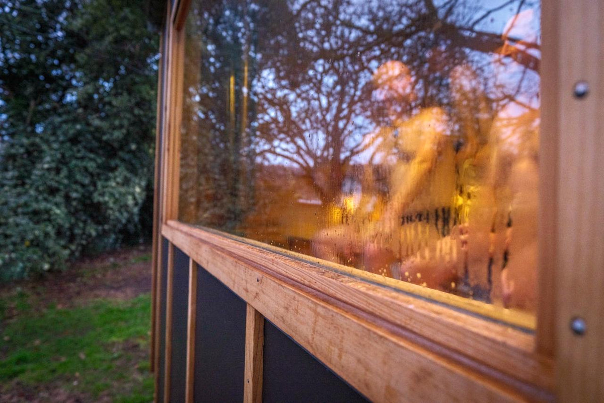 A close-up of a wooden-framed window with a glass pane, showing warm interior lighting and blurred reflections of trees and people outside in the evening.