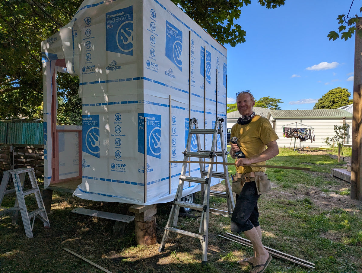 A man wearing a yellow t-shirt and shorts stands next to a custom-built outdoor shower enclosure wrapped in white insulation boards with blue logos. The enclosure is set in a backyard with grass, trees, and ladders.