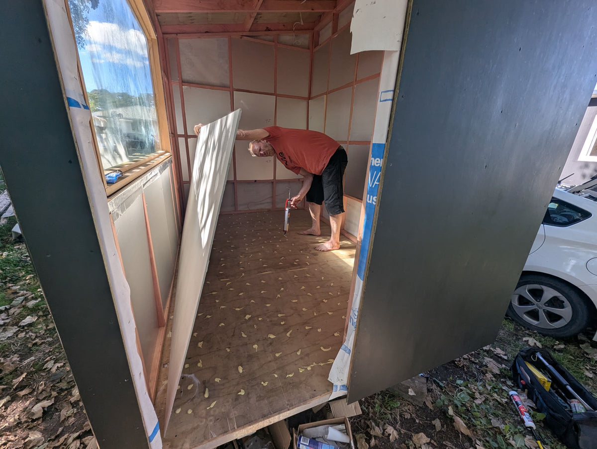 A man installs a large white panel inside a wooden shed or cabin structure, working on a dirt floor. The exterior of the structure shows dark gray siding, and a white car is parked outside on the right.