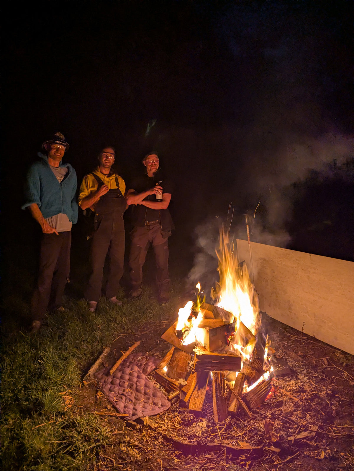 Three people standing outdoors at night beside a burning campfire, with grass, smoke, and a dark background visible.