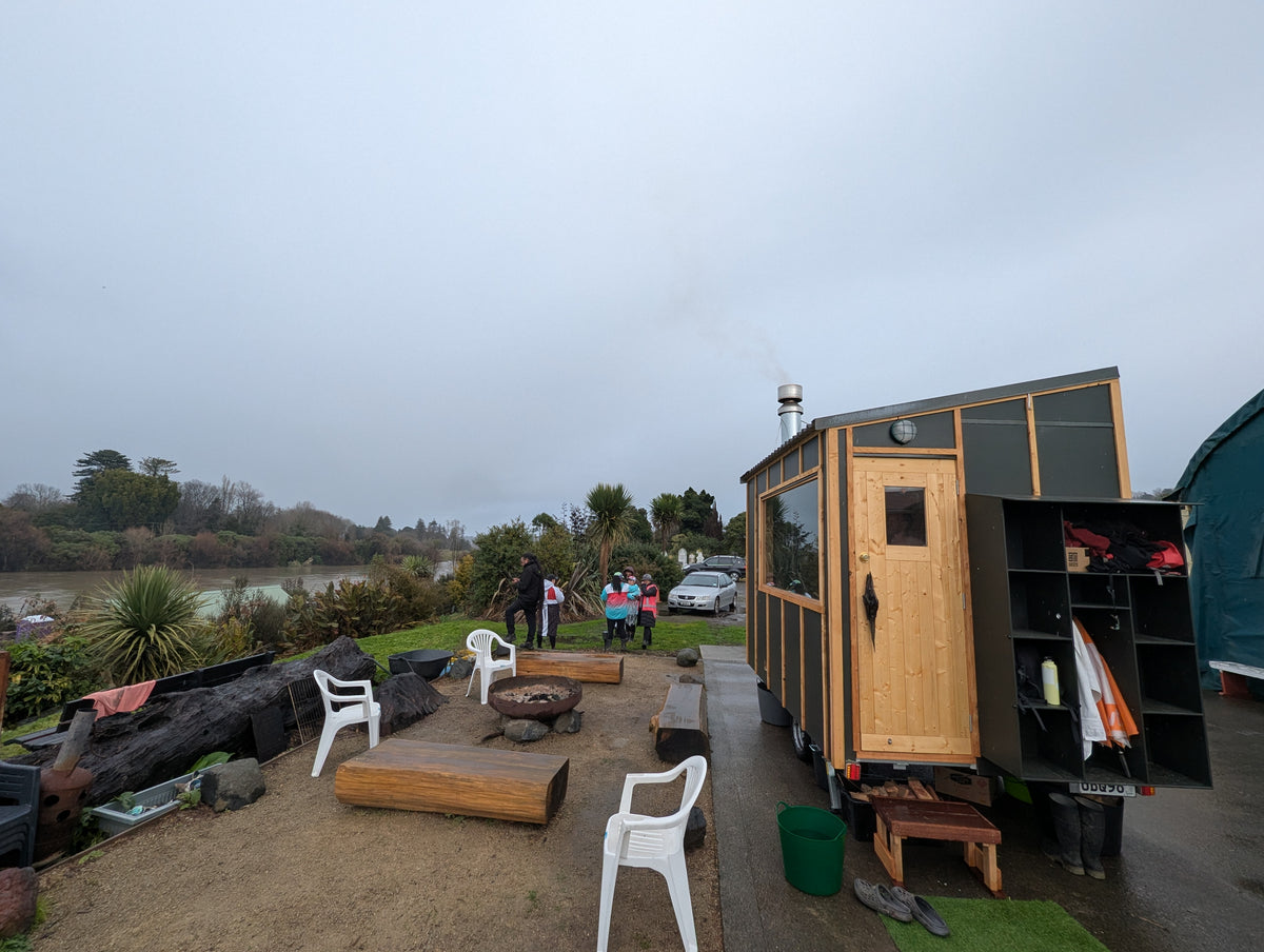 A small mobile sauna with a wood-grain door and dark green exterior is parked outdoors on a gravel pad next to the whanganui river. 